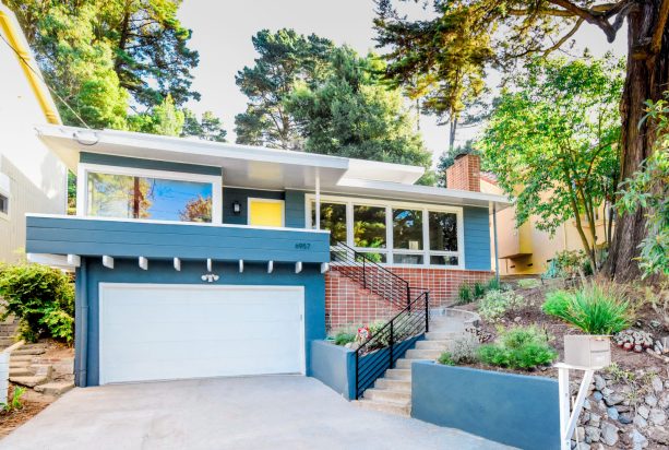 a 1960s blue exterior home with a yellow door combined with red bricks and a white roof
