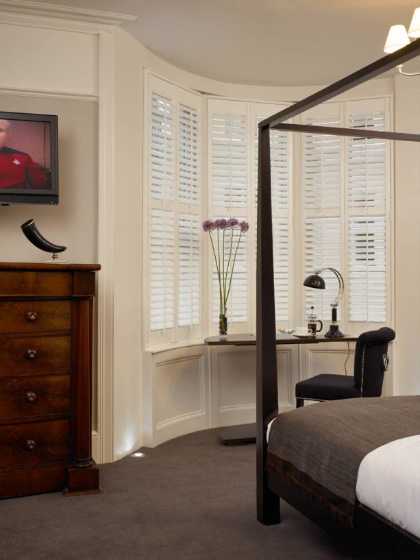a dark brown one leg wooden desk is perfect for a beige narrow bay window with plantation shutters