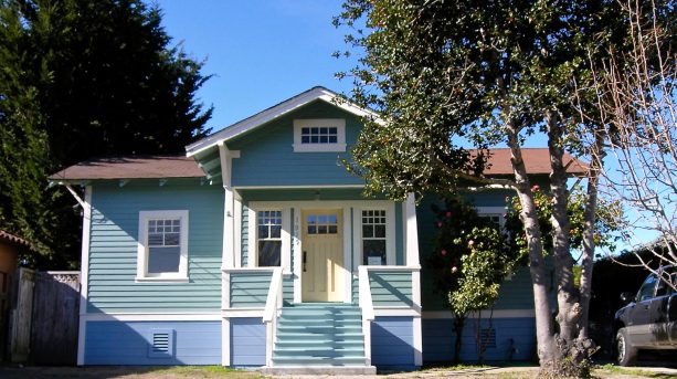 light blue small craftsman house with a yellow door and a brown roof