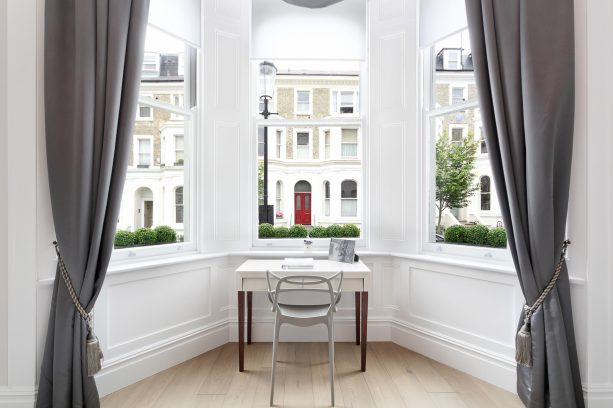 the contrasting sizes of a small white freestanding desk with brown legs and a big bay window add an interesting look to a transitional home office