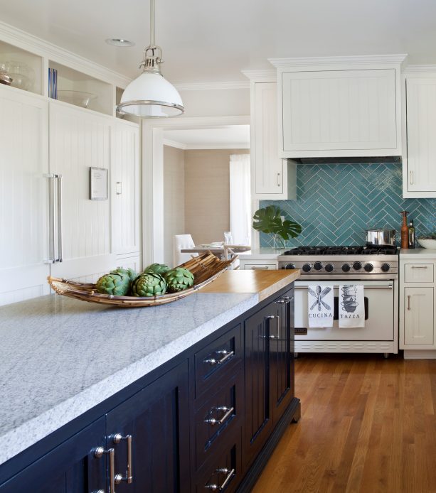 a kitchen with a navy island and white beaded inset cabinets accentuated by an aqua backsplash